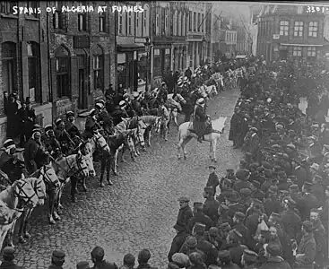 Photo noir et blanc montrant une trentaine de cavaliers sur un rang, portant burnous, au bord d'une place, face à la foule des civils.
