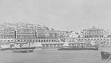 Vue du port d'Alger dans les années 1930. On voit la Méditerranée, quelques bateaux, des façades de bâtiments assez imposants, des arcades. Le reste de la ville se dévoile en hauteur en arrière-plan. La photo est en noir et blanc.