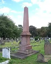 Un grand obélisque rouge à quatre côtés entourée de pierres tombales beaucoup plus petites.