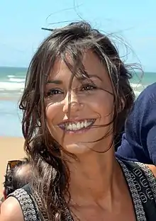 Portrait d'une jeune femme à la peau mate, arborant un large sourire. Sa chevelure brune est décoiffée par le vent. Au fond, on aperçoit une plage et un ciel bleu.