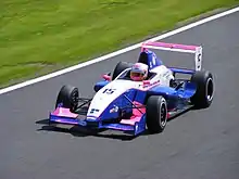 Photographie d'une monoplace de Formule Renault blanche, bleu foncé et rose, vue d'en haut, de trois-quart gauche, sur une piste sèche.