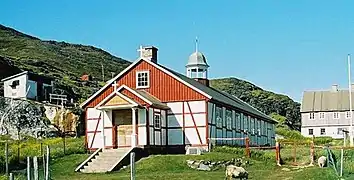 Photographie en couleurs d'un bâtiment en bois rouge et blanc entouré de poteaux de clôture et de parcelles herbacées, son toit surmonté d'un clocher, sa façade précédée d'un porche et d'un escalier.