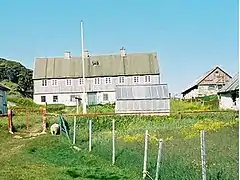 Photographie en couleurs de terrains herbus séparés d'une clôture grillagée, des bâtiments gris faits de tôles visibles au second plan.