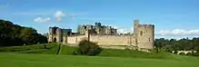Photographie donnant une vue d'ensemble du château d'Alnwick, avec ses longs remparts de pierre, dans la campagne anglaise verdoyante, par beau temps.