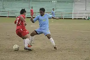 Photographie d'une scène d'un match de football : un joueur au maillot bleu ciel se dresse devant un joueur en rouge.