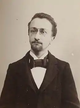 Photographie en noir et blanc d'un homme, en buste, portant un costume, un nœud papillon et des lunettes.