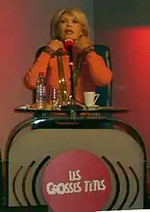 Photo d'Amanda Lear assise derrière un pupitre orné d'un micro et affichant le logo de l'émission Les grosses Têtes.