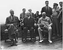 Photographie en noir  et blanc de trois hommes assis dans des fauteuils et discutant ; d'autres hommes sont debout derrière eux.
