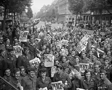 Foule de soldats alliés en liesse dans une rue ; plusieurs brandissent un journal avec le mot Piece inscrit en grand.