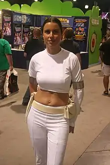 Femme en veste et pantalon blancs devant un stand d'exposition.