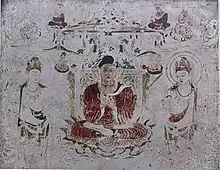 Photo couleur d'une fresque murale figurant, en son centre, un bouddha assis dans la position du lotus, entouré de deux disciples (teinte beige dominante).
