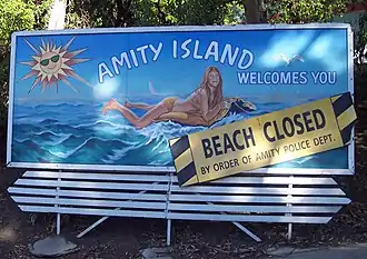 Large panneau représentant une jeune femme sur un matelas gonflable sur la mer, disant en anglais « Amity Island vous accueille » avec un rajout qui précise que la plage est fermé par ordre de la police