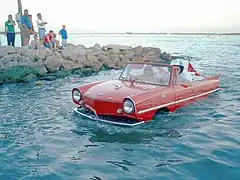 "véhicule rouge sortant de l’eau, avec de l’eau jusqu’au-dessus des roues, 4 personnes a bord, quelques spectateurs sur une jetée."
