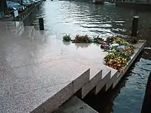 Photo en couleurs (teinte bleutée dominante), prise à Amsterdam en novembre 2003, et montrant le « Homomonument » : un monument dédié aux victimes homosexuelles du nazisme. La photo en montre seulement une partie : un assemblage de cinq dalles en granit rose qui forment un escalier triangulaire permettant d’accéder, du trottoir, au bord d’un canal (de gauche à droite sur la photo). La dernière marche, une plate-forme triangulaire, qui semble flotter sur l’eau du canal, est en partie recouverte de gerbes de fleurs. L’arrière-plan de la photo est constitué par l’étendue d’eau du canal.