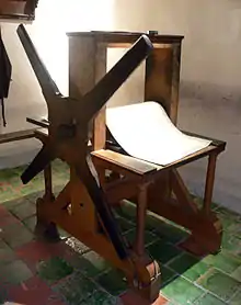 Photographie en couleurs. Une presse en bois constituée d'une grande manivelle à quatre bras est éclairée dans une pièce fermée. Sur le rouleau, un feutre blanc attend d'être déposé sur une plaque à imprimer.