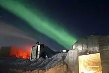 Des bâtiments métalliques dans un paysage enneigé, dans la nuit. Une aurore boréale verte et des étoiles sont visibles dans le ciel.