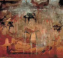 Photographie d'une peinture murale, sur laquelle on voit au centre, assis en tailleur, une personne habillée en costume ancien, ainsi que deux autres personnes de plus petite taille lui ressemblant.