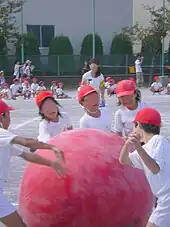 Des enfants vêtus de blanc et portant des casquettes rouges poussent une grosse balle rouge.