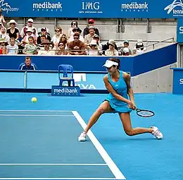 Une joueuse de tennis en robe bleue claire plie le genou pour taper une balle basse d'un revers à deux mains.