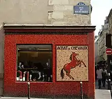 Devanture d'un magasin de briques rouges sur lequel est écrit « achat de chevaux », un cheval étant représenté.