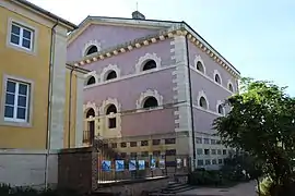 Photographie en couleurs d'un bâtiment à étages en maçonnerie rose éclairé de petites baies en demi-cercle.