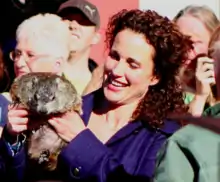 Phographie d'une femme portant une chemise violette. Elle se trouve au milieu d'une foule et tient dans ses mains une marmotte.