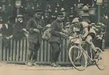 Cycliste entrant sur la piste d'un vélodrome sous le regard de deux agents et de la foule dans les tribunes.