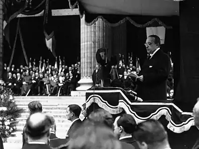 Photo en noir et blanc d'un homme prononçant un discours depuis une tribune, à proximité des marches d'un bâtiment à colonnes.
