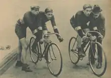Deux cyclistes prêts pour le départ, tenus par deux hommes debout.