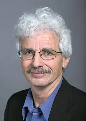 Portrait d'un homme aux cheveux blancs-gris, portant des lunettes et une moustache grise.