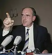 Photographie d'un homme en costume pointant du doigt devant un micro.