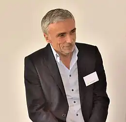 Photographie en couleurs d'un homme grisonnant et souriant, portant une veste noire, légèrement tourné à gauche.