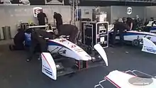 Photo d'un stand de course, où deux mécaniciens travaillent sur le cockpit d'une monoplace