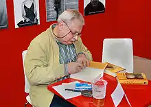 Photographie couleur d'un homme vieillissant, assis à une table et en train de dédicacer des livres.