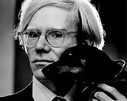 Portrait photographique en noir et blanc d'un homme blond portant des lunettes tenant dans sa main un petit chien