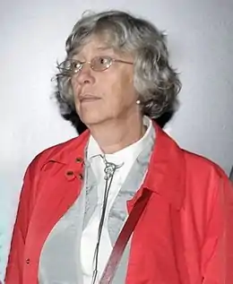 Portrait d'une femme d'une soixantaine d'années, avec des cheveux gris, des lunettes de vue et une veste rouge.