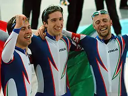 Photographie de trois patineurs en combinaison blanche et bleue, les bras levés, enveloppés d'un drapeau italien.