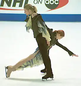 Photographie de deux patineurs sur une patinoire, un homme debout, une femme penchée en avant se retenant à lui.