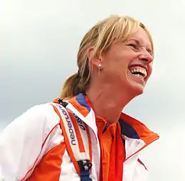 Une femme blonde coiffée d'une queue de cheval et portant une veste de sport blanche et orange rit aux éclats.