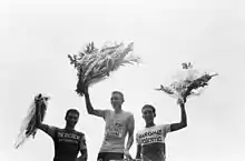 Photographie en noire et en blanc montrant trois cyclistes tenant un bouquet de fleurs sur le podium après l'arrivée d'une course.