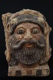 Antéfix (motif initialement placé sur un toit ou une corniche) en terre-cuite peint en noir et en rouge représentant la tête d'un homme souriant avec une barbe et une moustache.