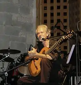 Photographie couleur d'un guitariste assis, parlant au micro en tenant son instrument.