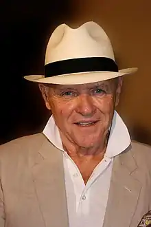 Portrait de Hopkins souriant sur un fond sombre. Il porte un costume clair, un polo blanc et un chapeau.