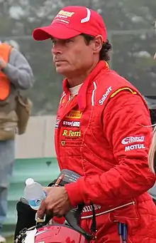 Photographie d'un homme blanc, brun, de trois-quarts gauche, debout, en combinaison rouge et casquette rouge, en plan américain.