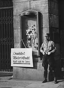 Photographie en noir et blanc d'un  membre de la SA à côté d'une affiche proclamant : « Allemands ! Défendez-vous ! N'achetez pas chez les Juifs ! » en 1933
