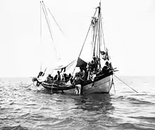 Cliché noir et blanc. Un scaphandrier sur le flanc d'un bateau. Un autre, sans casque de plongée, à cheval sur le bastingage. Sept hommes d'équipage d'un bateau à voile ancré.