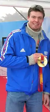 Photographie d'un homme en jean et veste bleue portant l'inscription France et les anneaux olympiques, avec une médaille d'or autour du cou qu'il tient de sa main droite.
