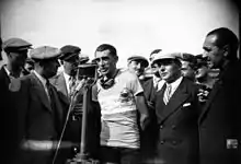 Photographie en noir et blanc montrant un cycliste parlant dans un micro, entouré par plusieurs personnes.