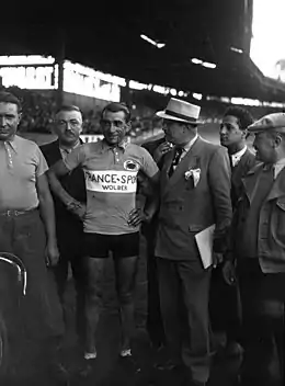 Photographie en noir et blanc montrant un coureur debout à côté de son vélo, entouré par des officiels.