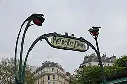 Photographie d'une arche en fer forgé surmontée d'une inscription sur laquelle deux pigeons semblent se pencher, en caractères stylisés de couleur gris foncé et en plus petits caractères rouges un nom suivi d'une abréviation